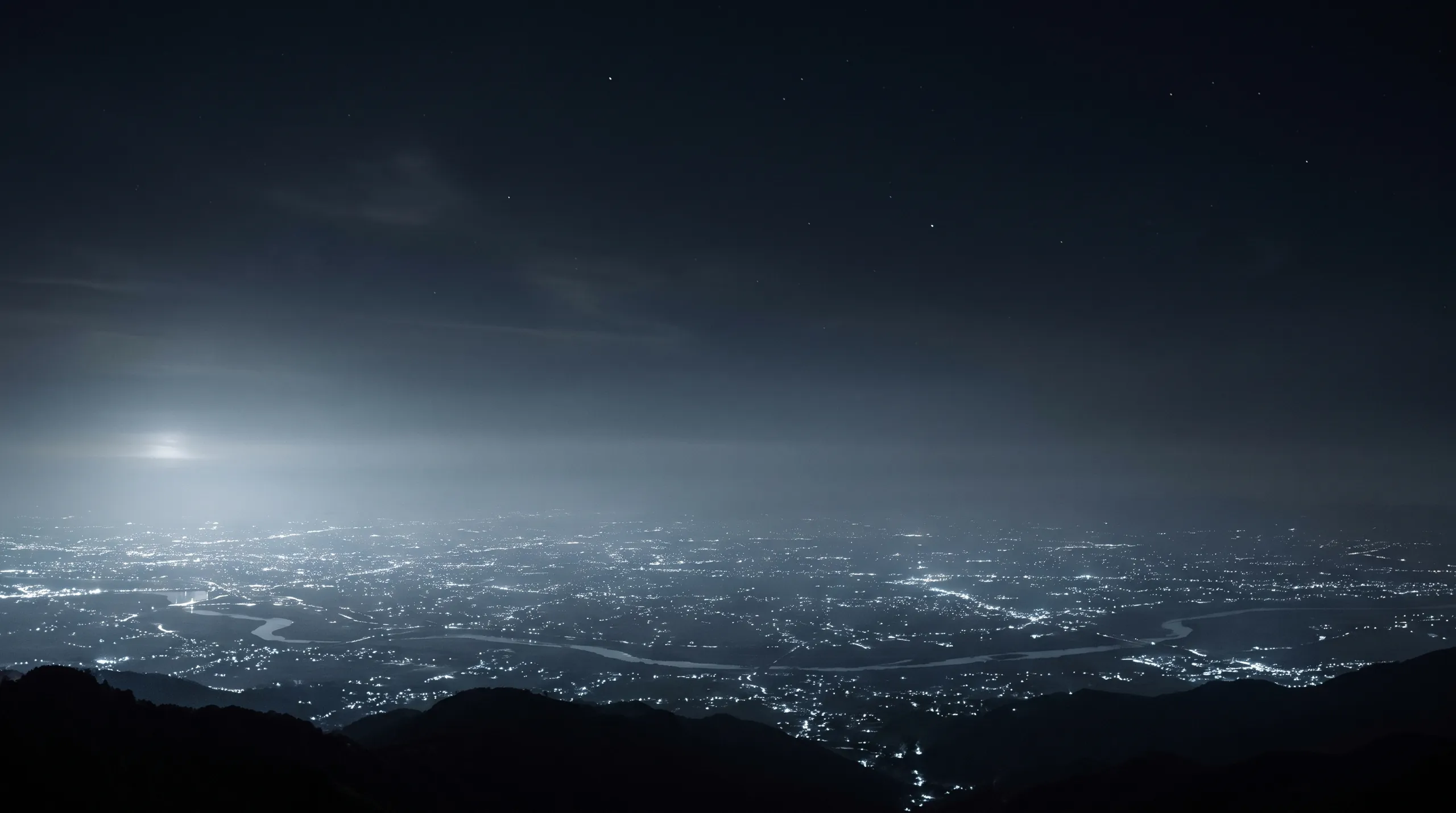 A valley city at night seen from a dark ridge, its lights spread under haze